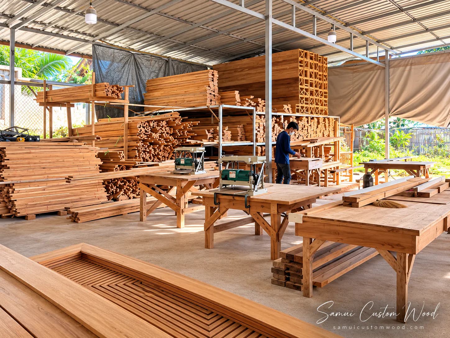 Inside the workshop: trestle benches with planers and racks of sawn teak, a carpenter working at the far bench