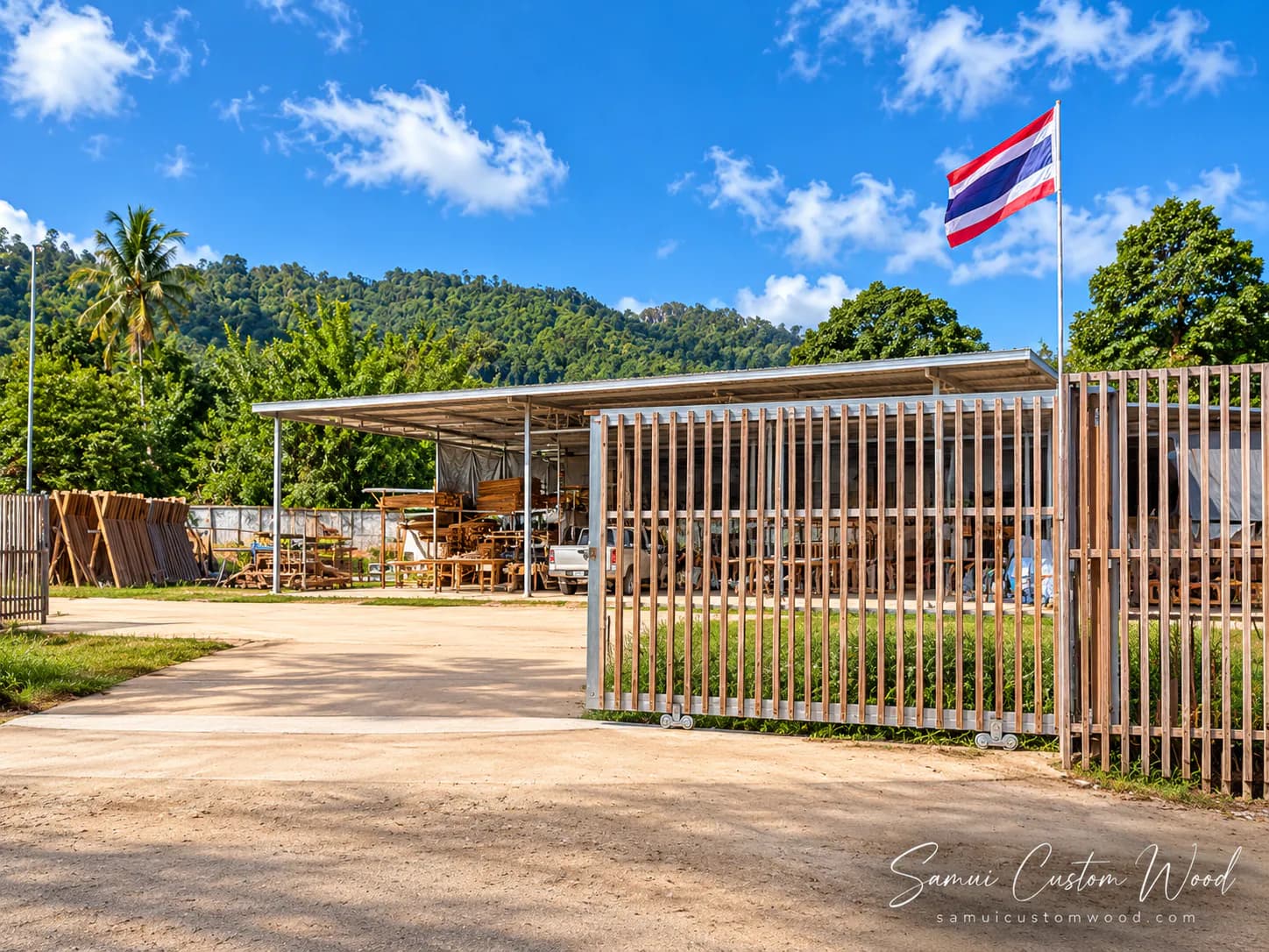 The workshop compound from the drive: a slatted gate, open-sided sheds stacked with timber and furniture, hills behind